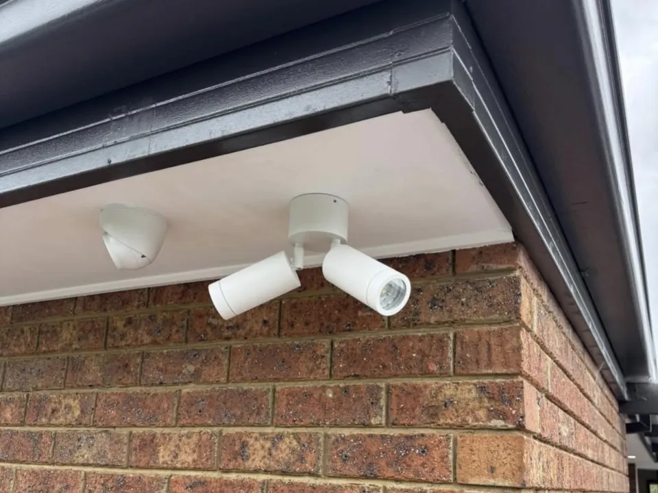Turret camera fitted beside twin adjustable spotlights under the eave of a brick home